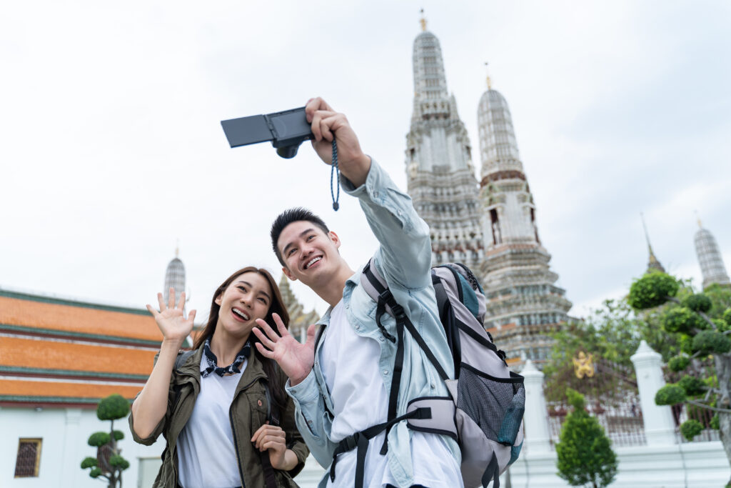 Asian couple backpacker using camera selfie and take a picture in city. Attractive man and woman tourist traveler travel on street, using smartphone record vlog on holiday vacation trip in Thailand.
